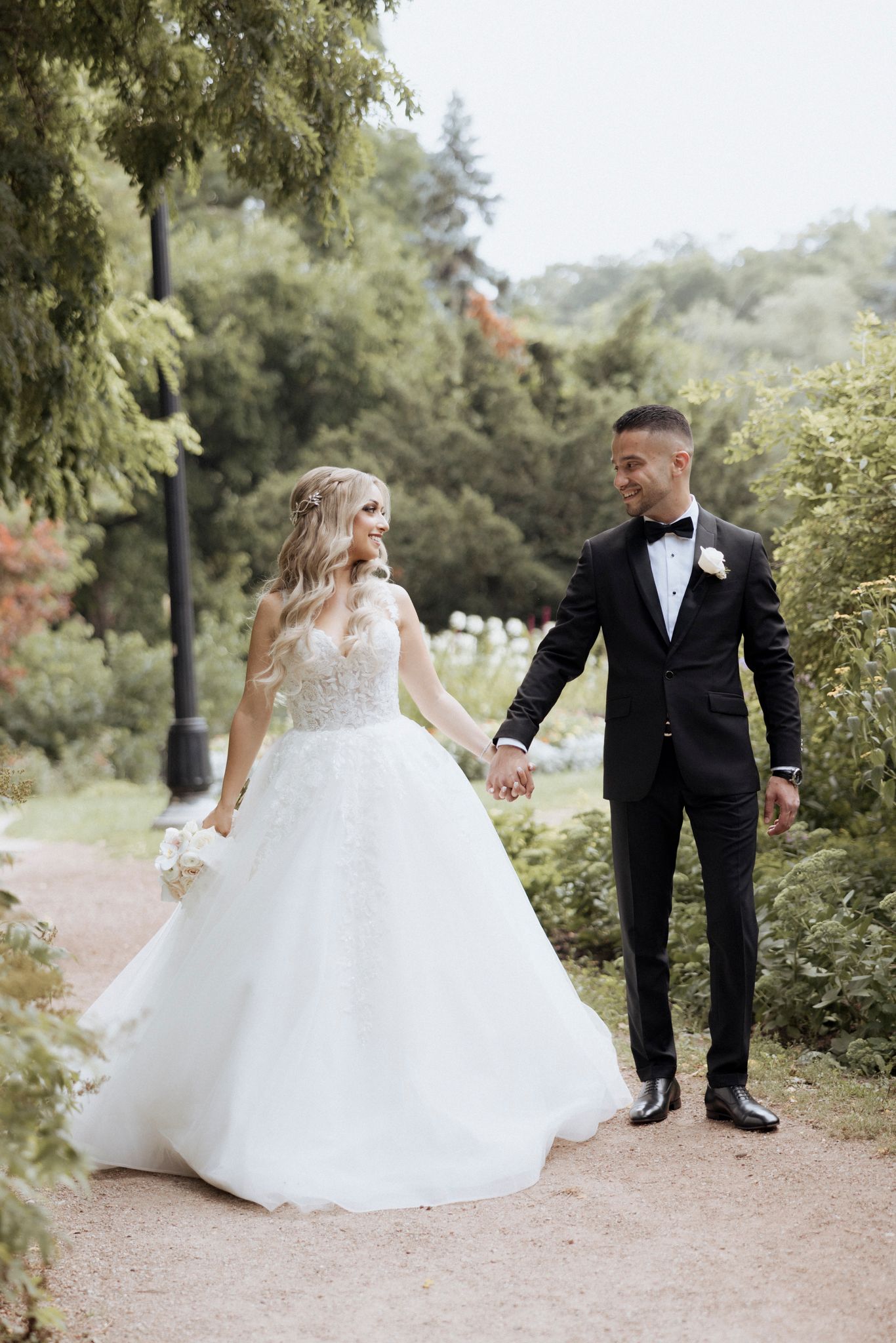 Newlyweds in formal attire holding hands while walking along garden path.
