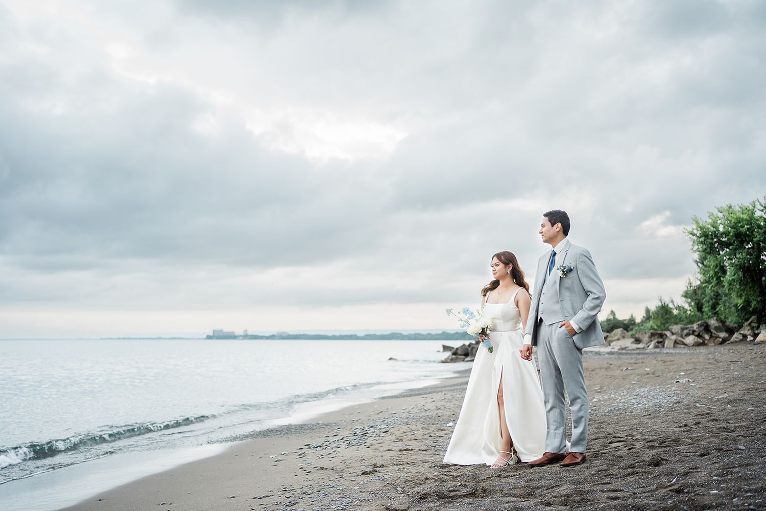 A couple strolls along a moody beach shoreline under cloudy skies wearing wedding attire.