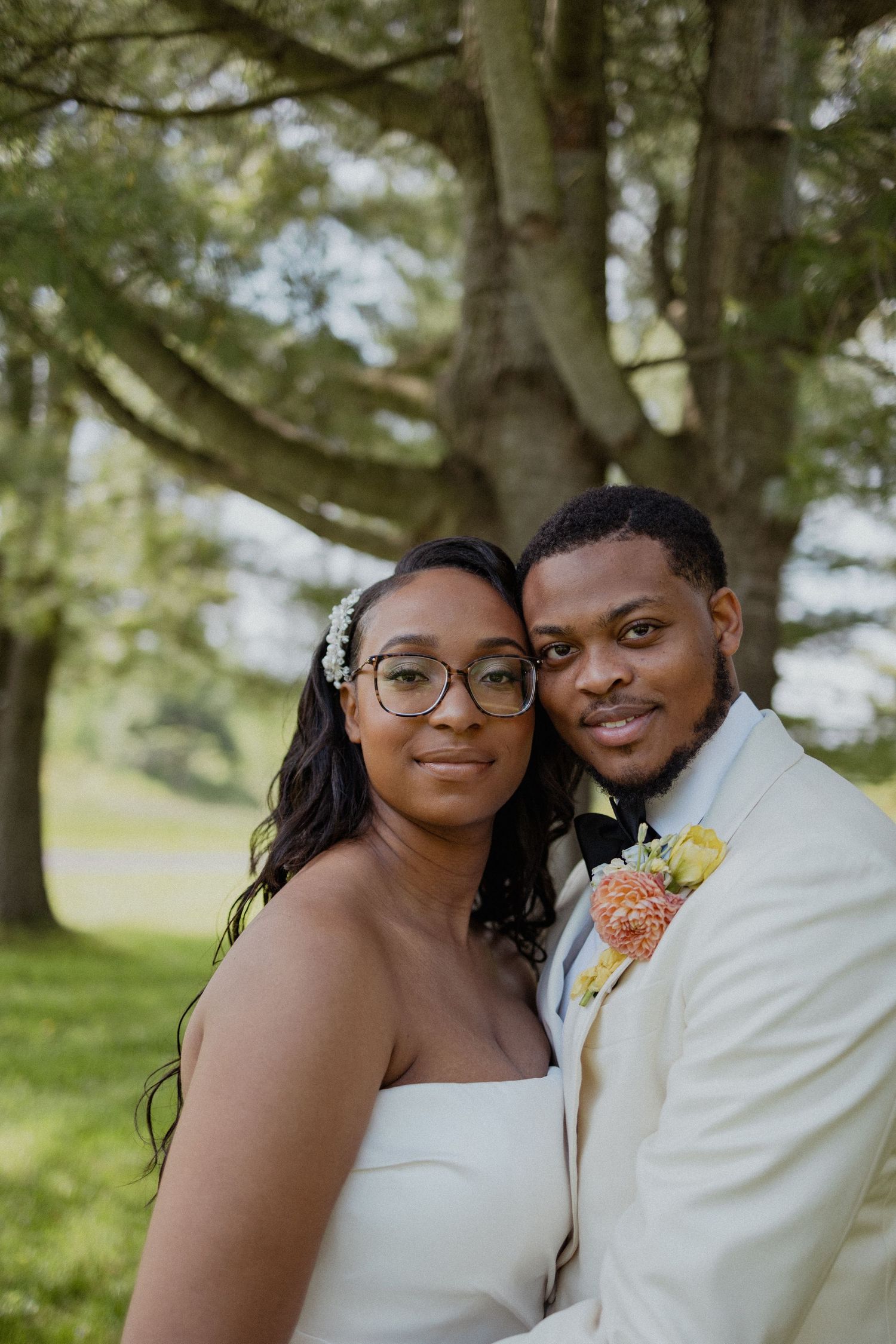 A romantic wedding portrait of a couple in white attire posing outdoors under a leafy tree.