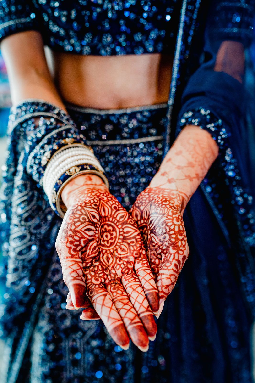Detailed henna designs adorn hands wearing traditional Indian bangles against a blue fabric background.