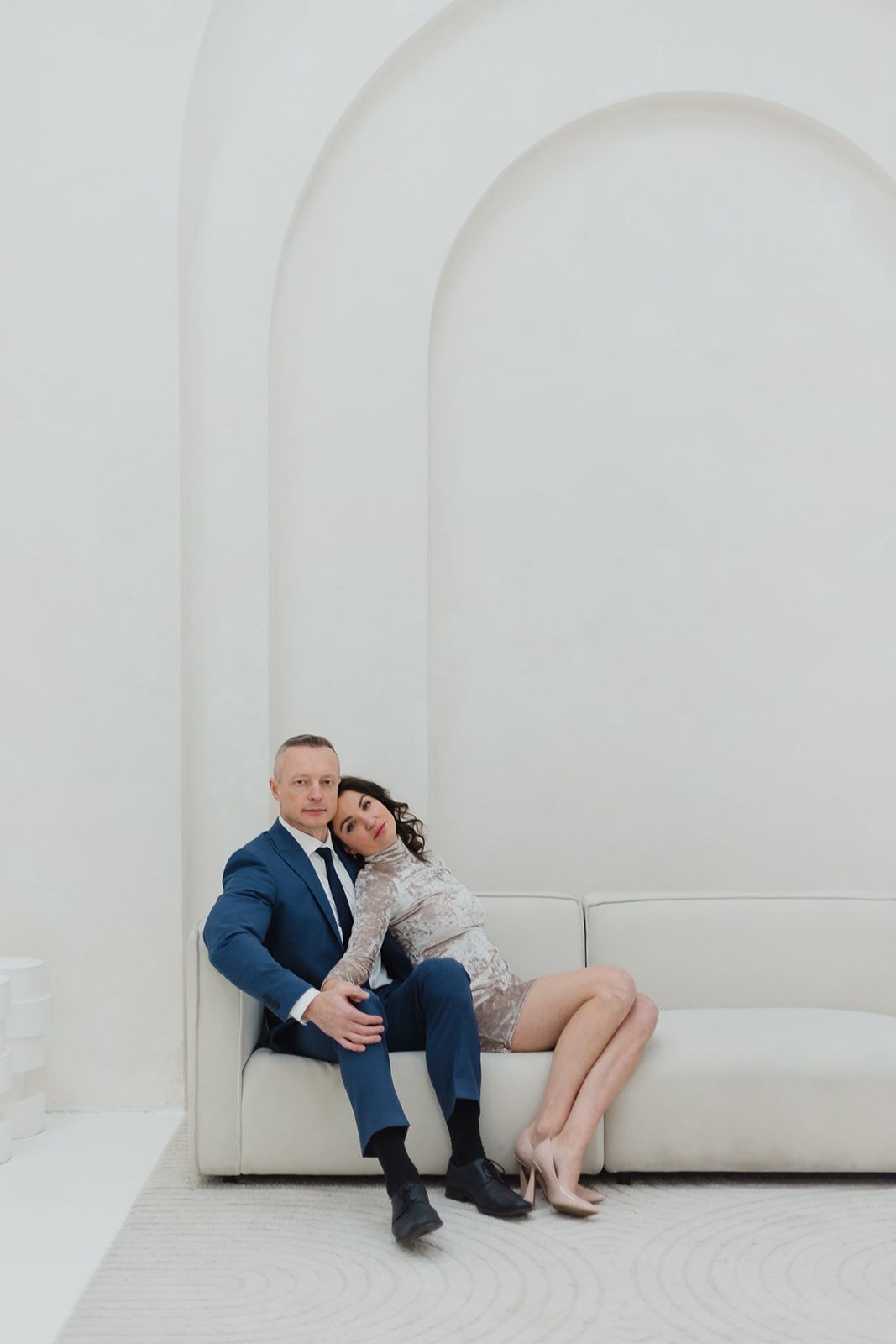 A couple in formal attire sits close together on a white modern couch against a minimalist white curved wall.