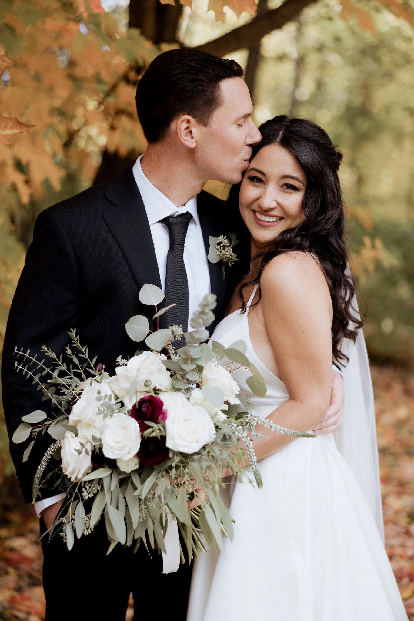 Wedding couple shares joyful moment in autumn setting with white and burgundy floral bouquet.