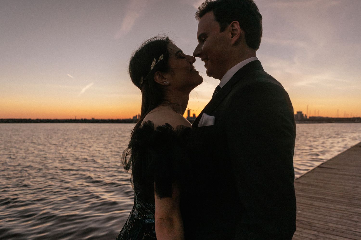 A silhouetted couple embraces on a wooden dock during sunset over calm waters.