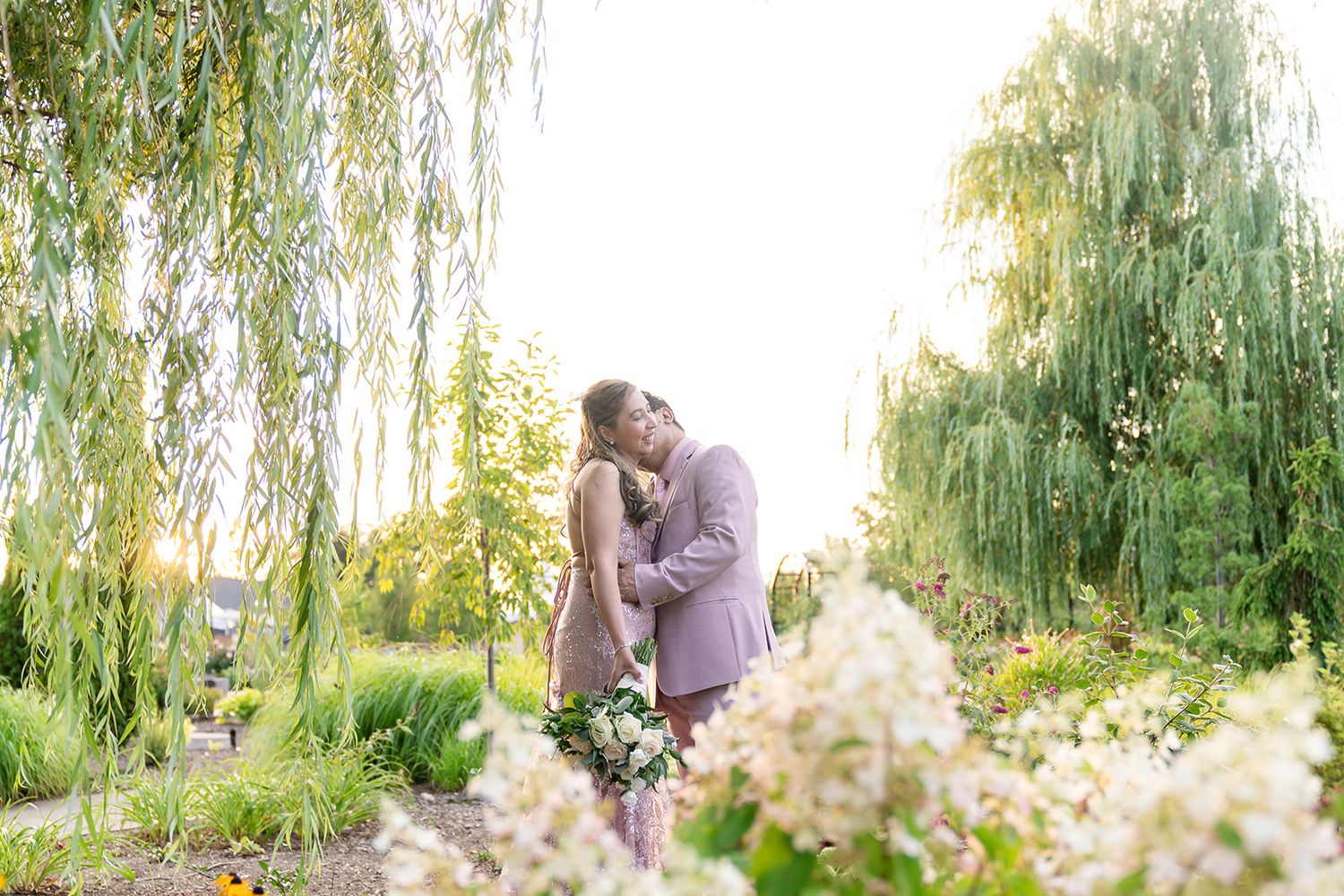 A romantic couple shares a tender kiss among blooming flowers and weeping willow trees in a garden at sunset.