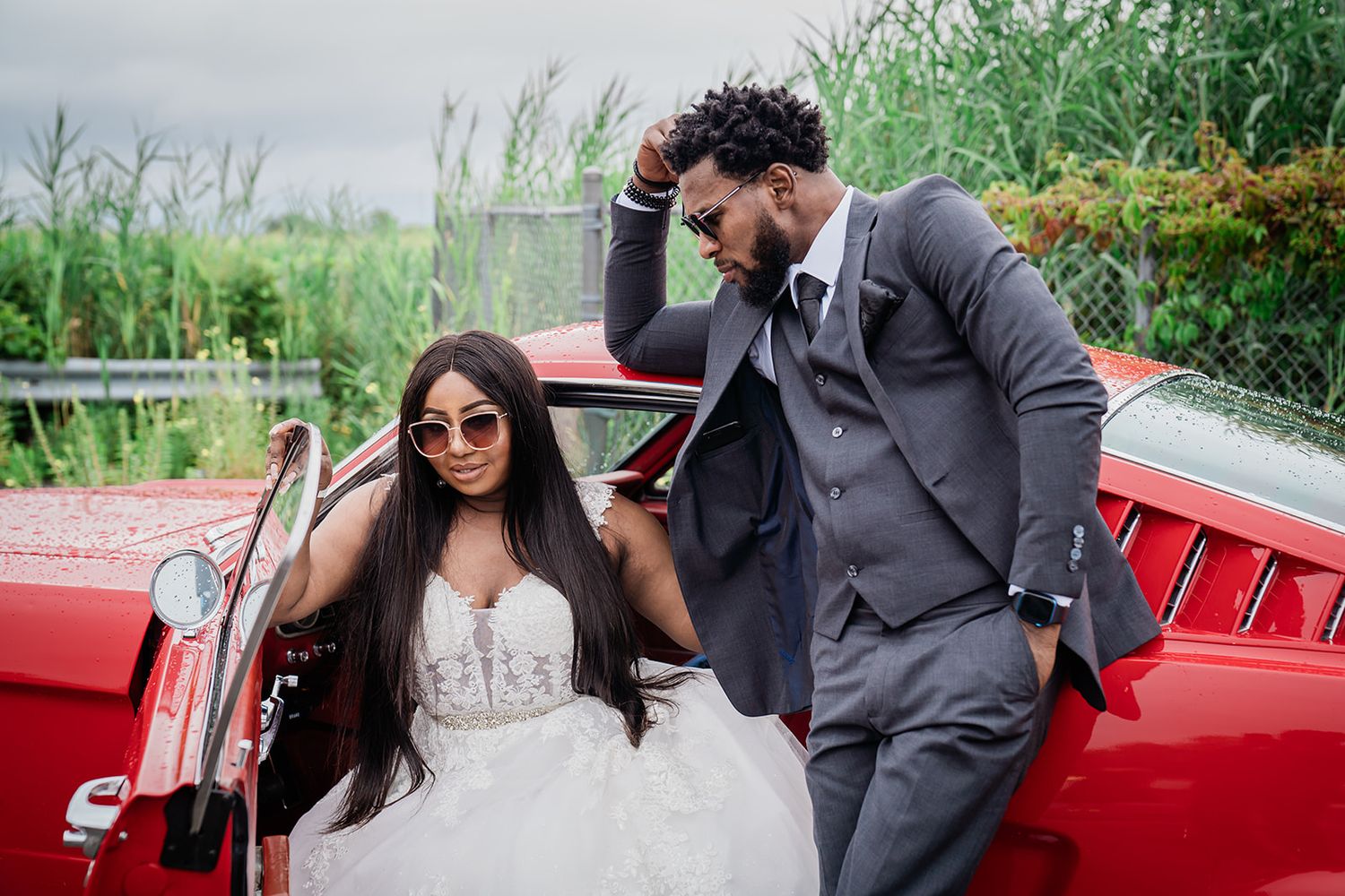 Wedding couple posing playfully with red classic car in garden setting.
