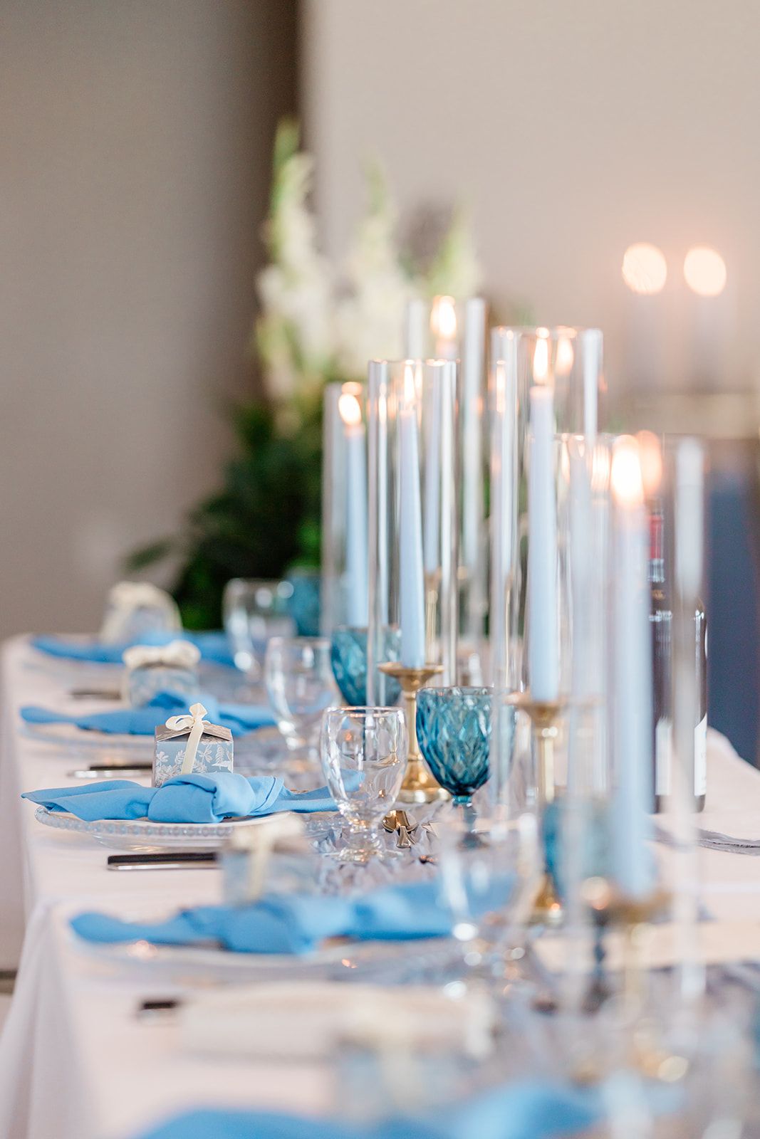 Elegant wedding table setting with white taper candles, blue napkins, and crystal glassware on a white tablecloth.