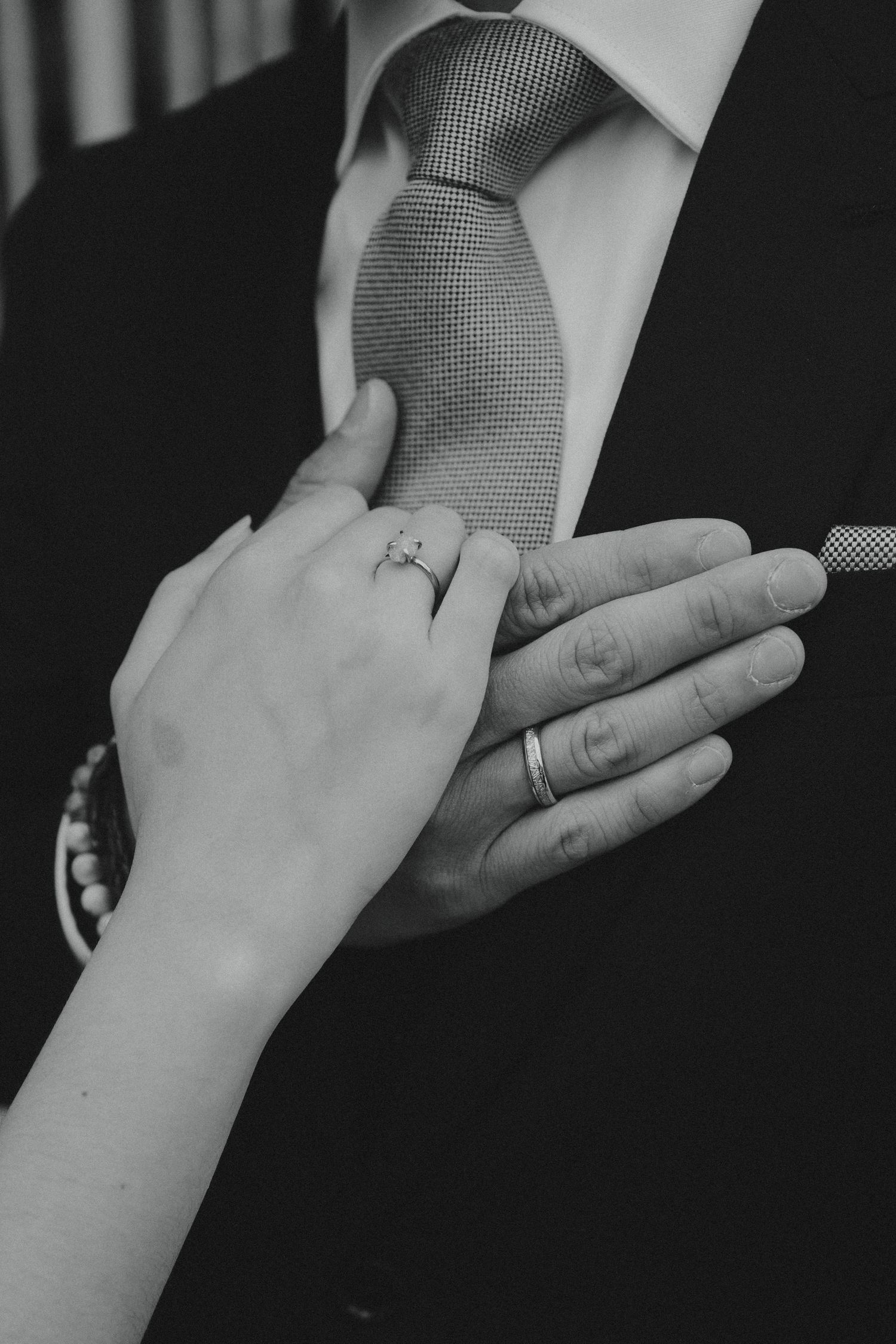 Black and white close-up of hands adjusting a textured tie, showing wedding rings on both hands.
