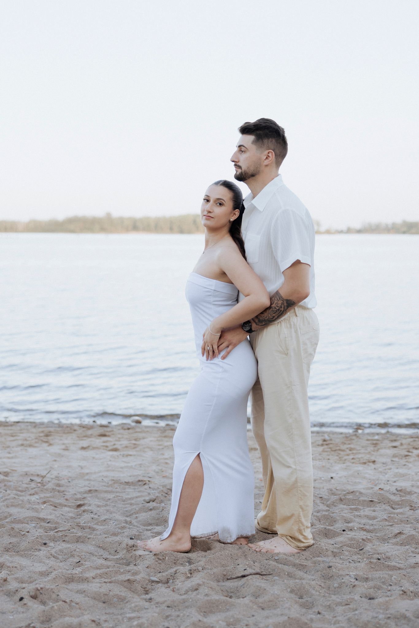 A couple in white outfits embraces on a sandy beach at sunset.