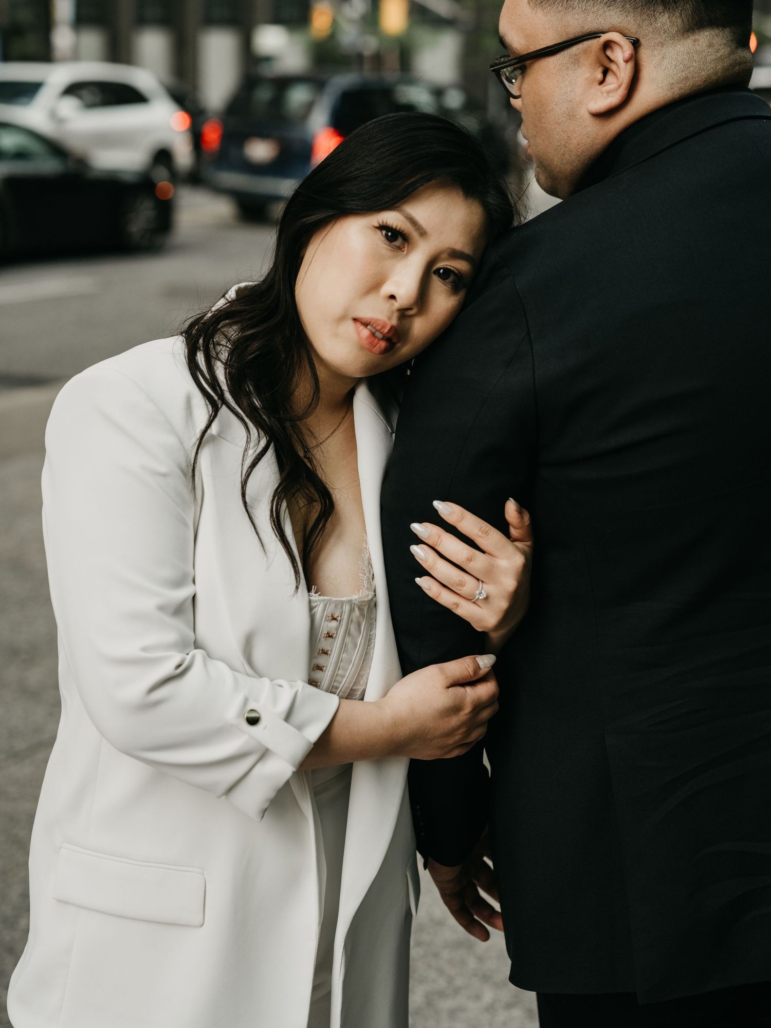 A couple shares an intimate moment on a city street as they embrace, with the woman wearing a white blazer.