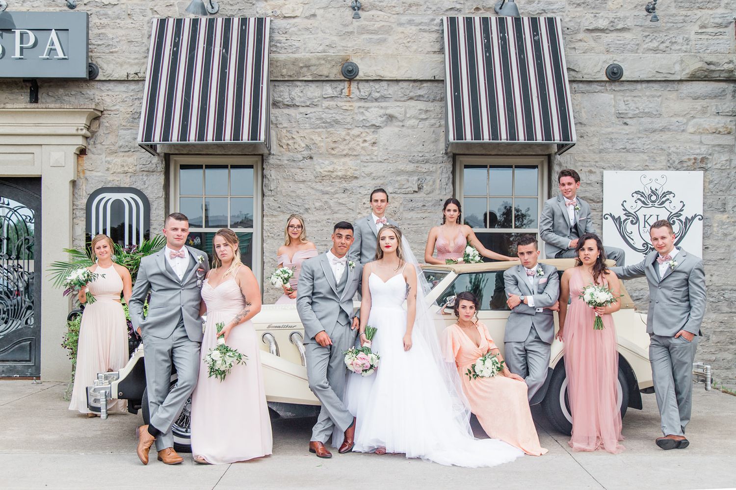 Wedding party in gray and blush attire poses together against stone wall with striped awnings.
