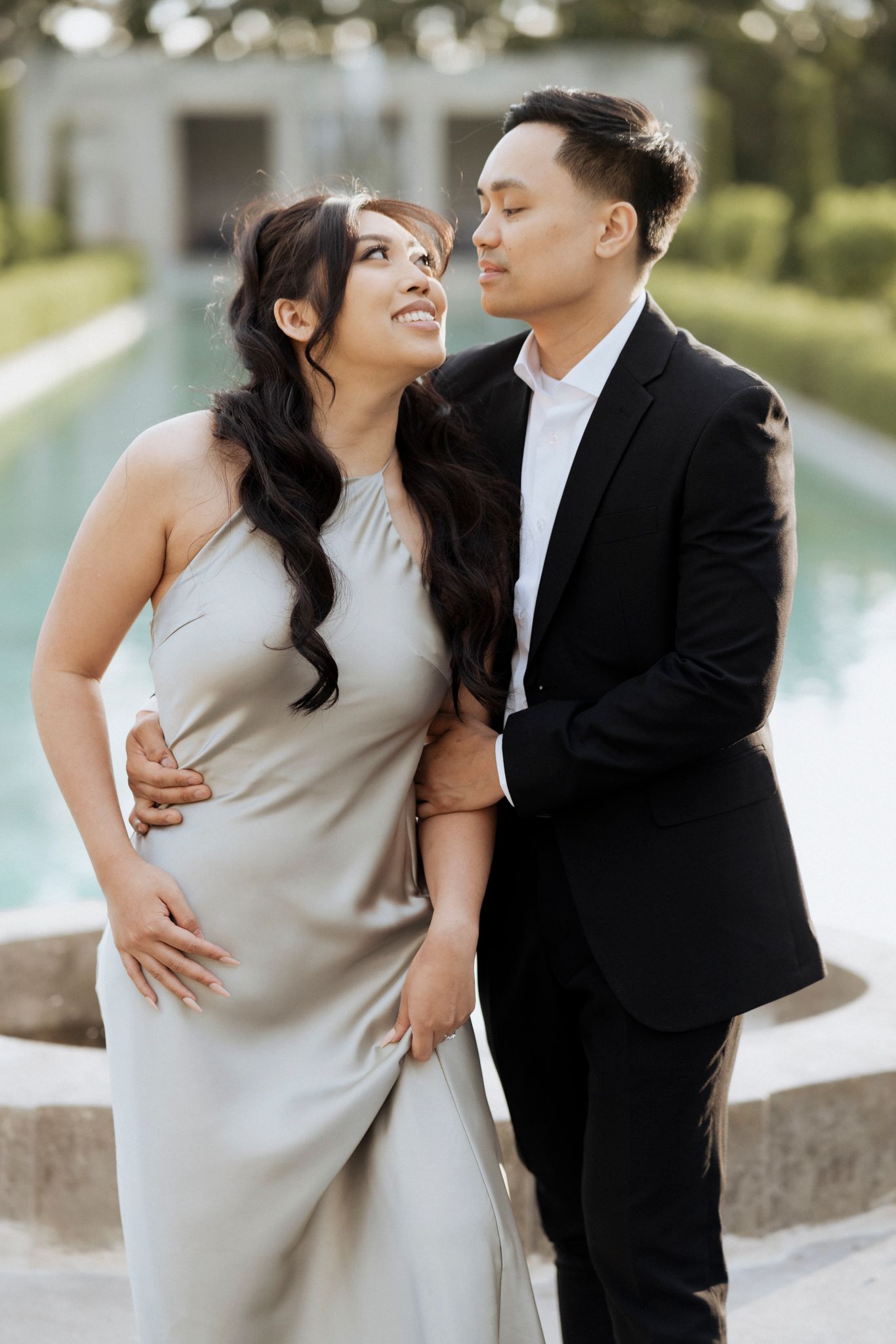 A couple in formal attire poses together by a pool during an elegant outdoor photoshoot.