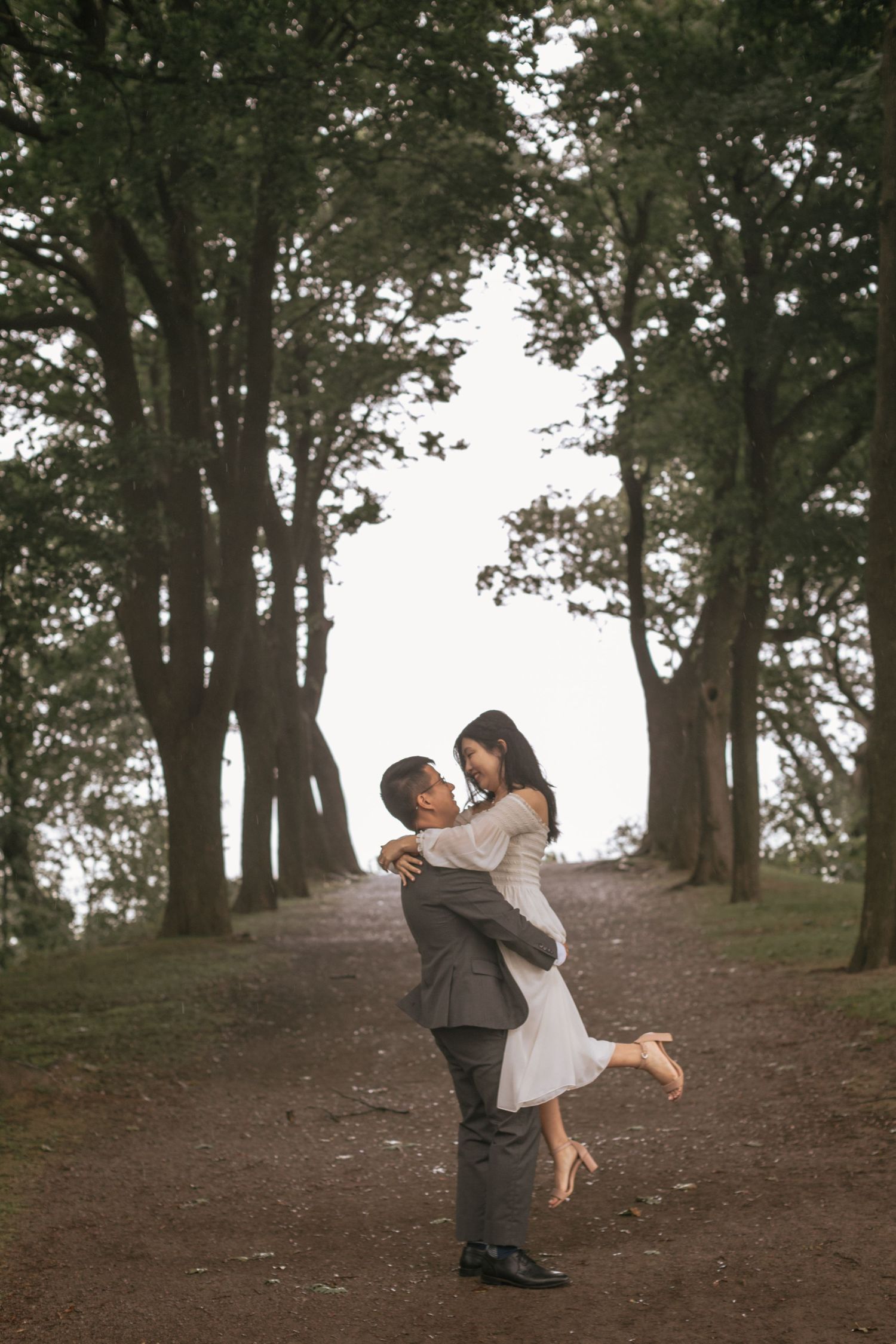 A romantic scene on a tree-lined path shows a couple embracing, with one person lifting the other in a joyful moment.