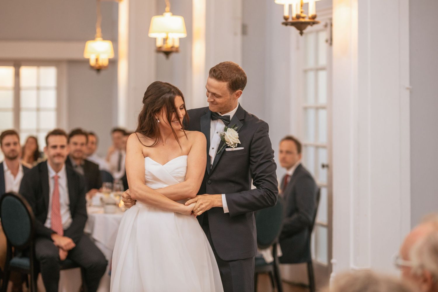 Newlywed couple shares an intimate moment during their first dance in an elegant ballroom with chandeliers.