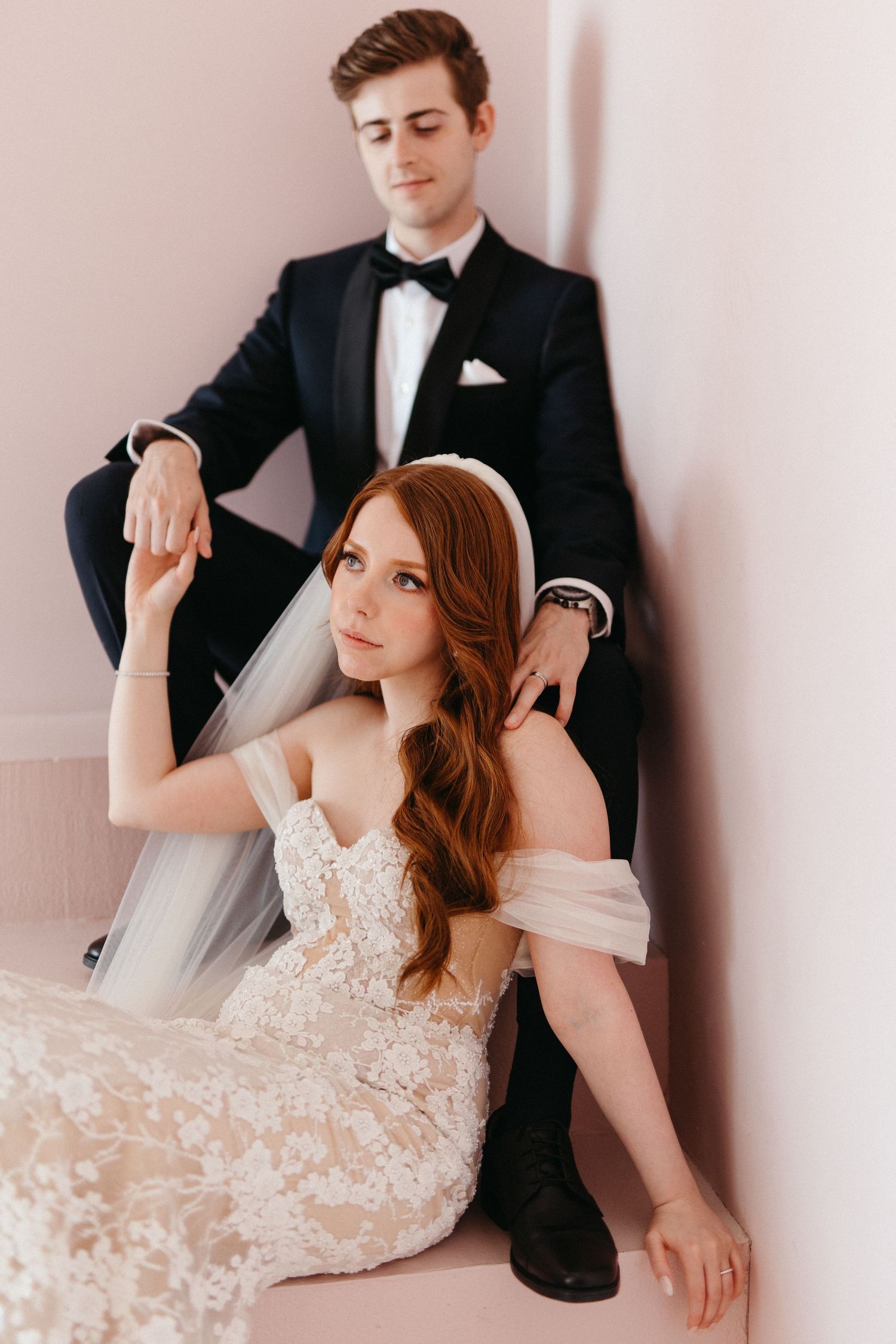 A couple in formal wedding attire pose together against a light wall in an elegant portrait session.