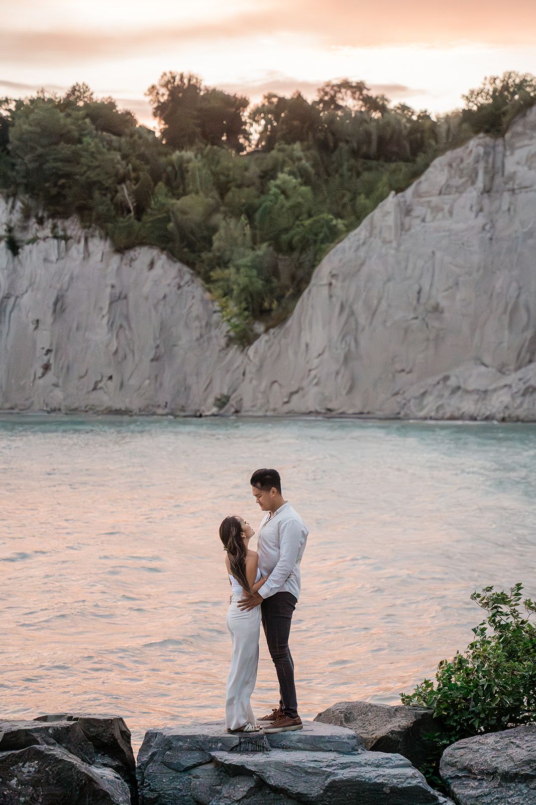 Couple standing on rocky shoreline at sunset against dramatic white cliffs.