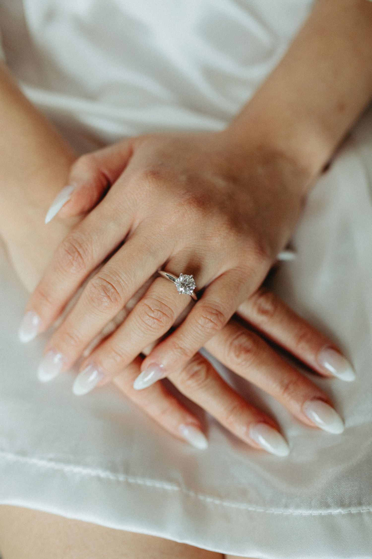 Close-up of manicured hands with white French tips and a sparkling diamond engagement ring on white fabric.