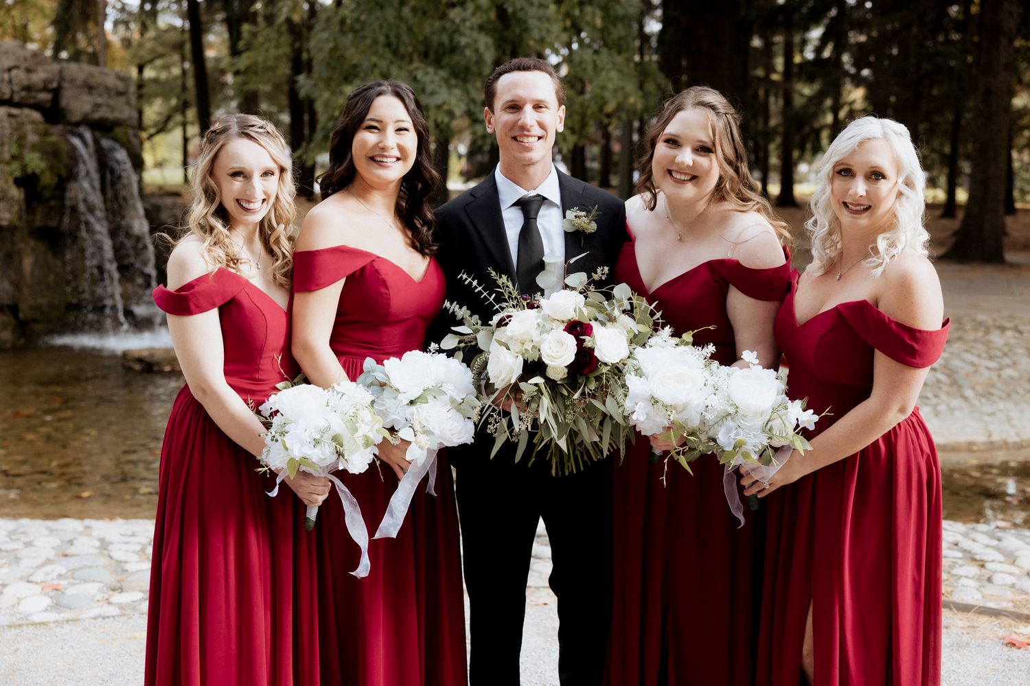 Wedding party in red dresses and formal attire posing with white floral bouquets.