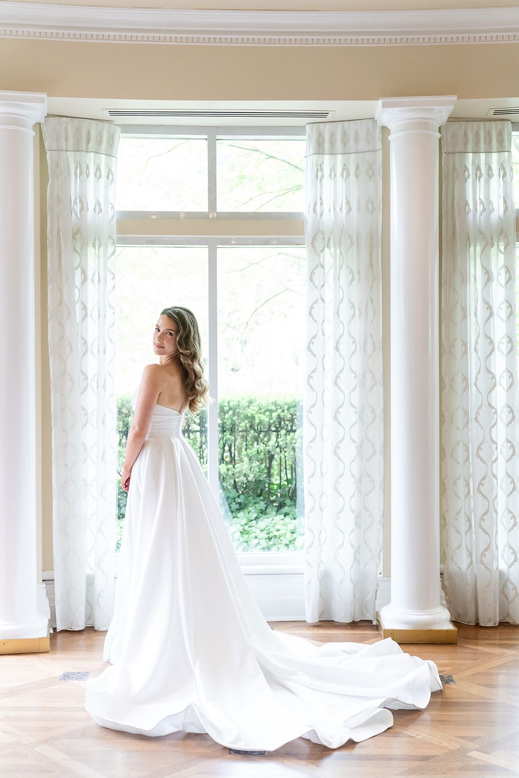 Elegant wedding gown with flowing train photographed in a bright sunlit room with white sheer curtains and columns.