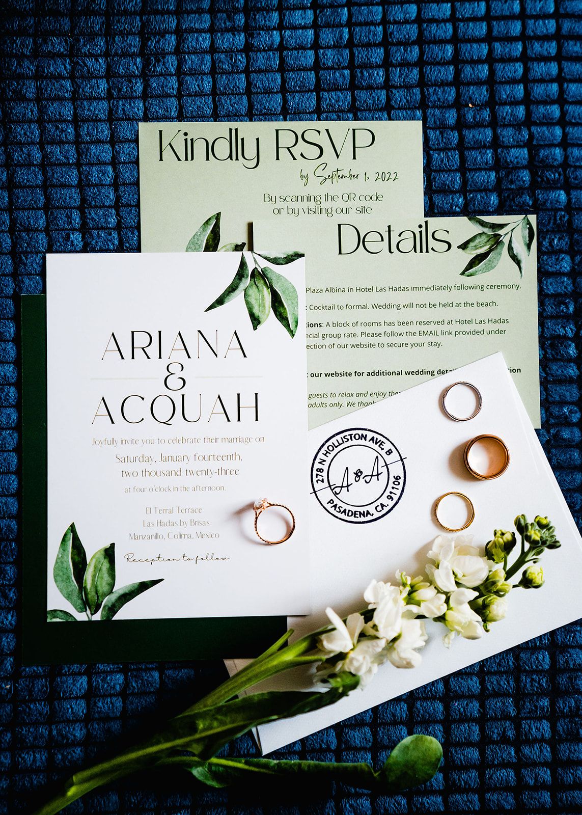 Wedding invitation with olive branch design and gold rings displayed with white flowers on a blue textured background.
