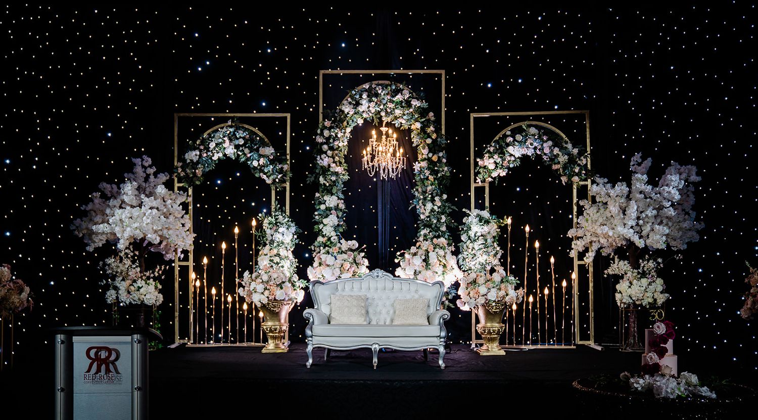 Elegant wedding stage decorated with white floral arrangements, golden archways and dramatic black backdrop illuminated by soft lights.
