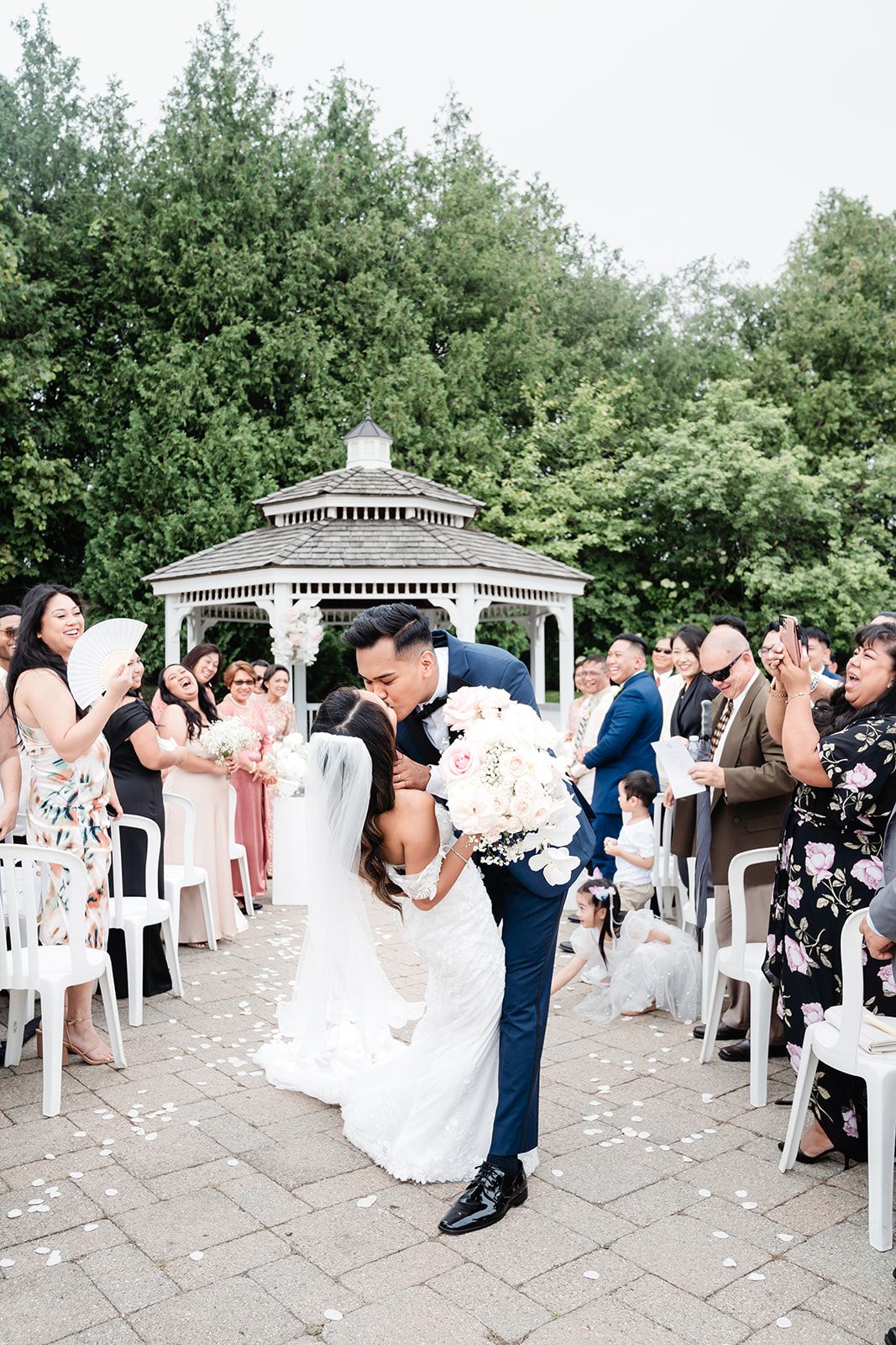 A romantic wedding ceremony takes place under a white gazebo surrounded by green trees and guests.