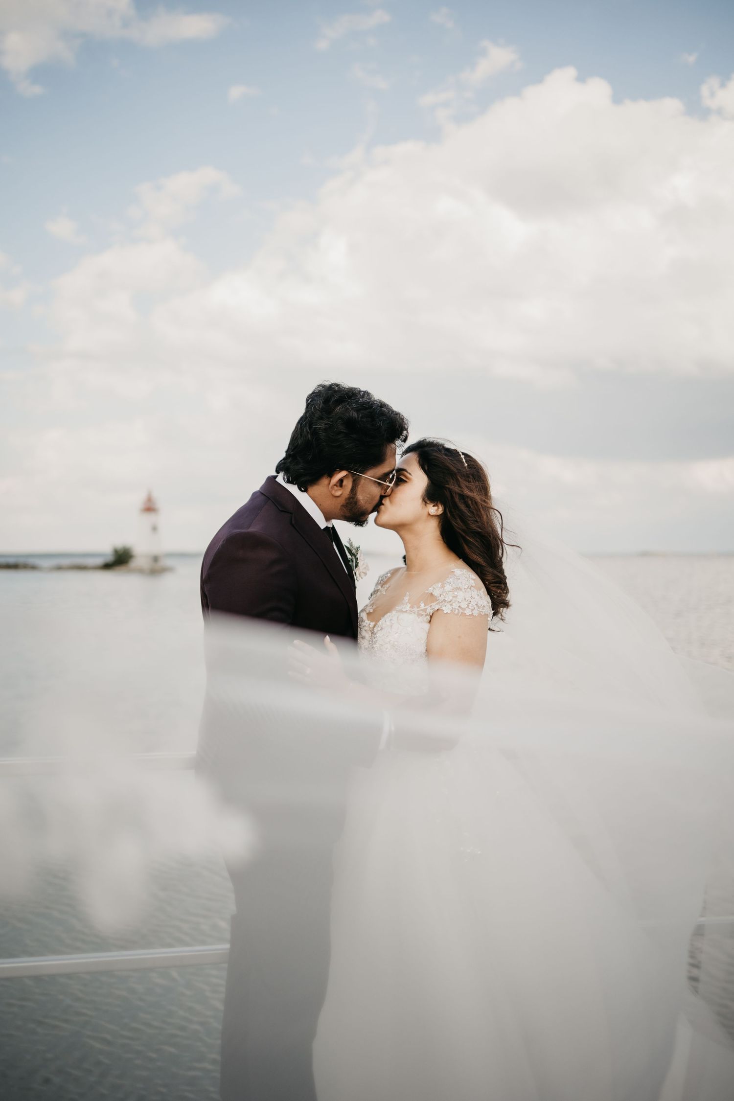 A romantic waterfront wedding photo with a lighthouse in the background and flowing veil in the foreground.