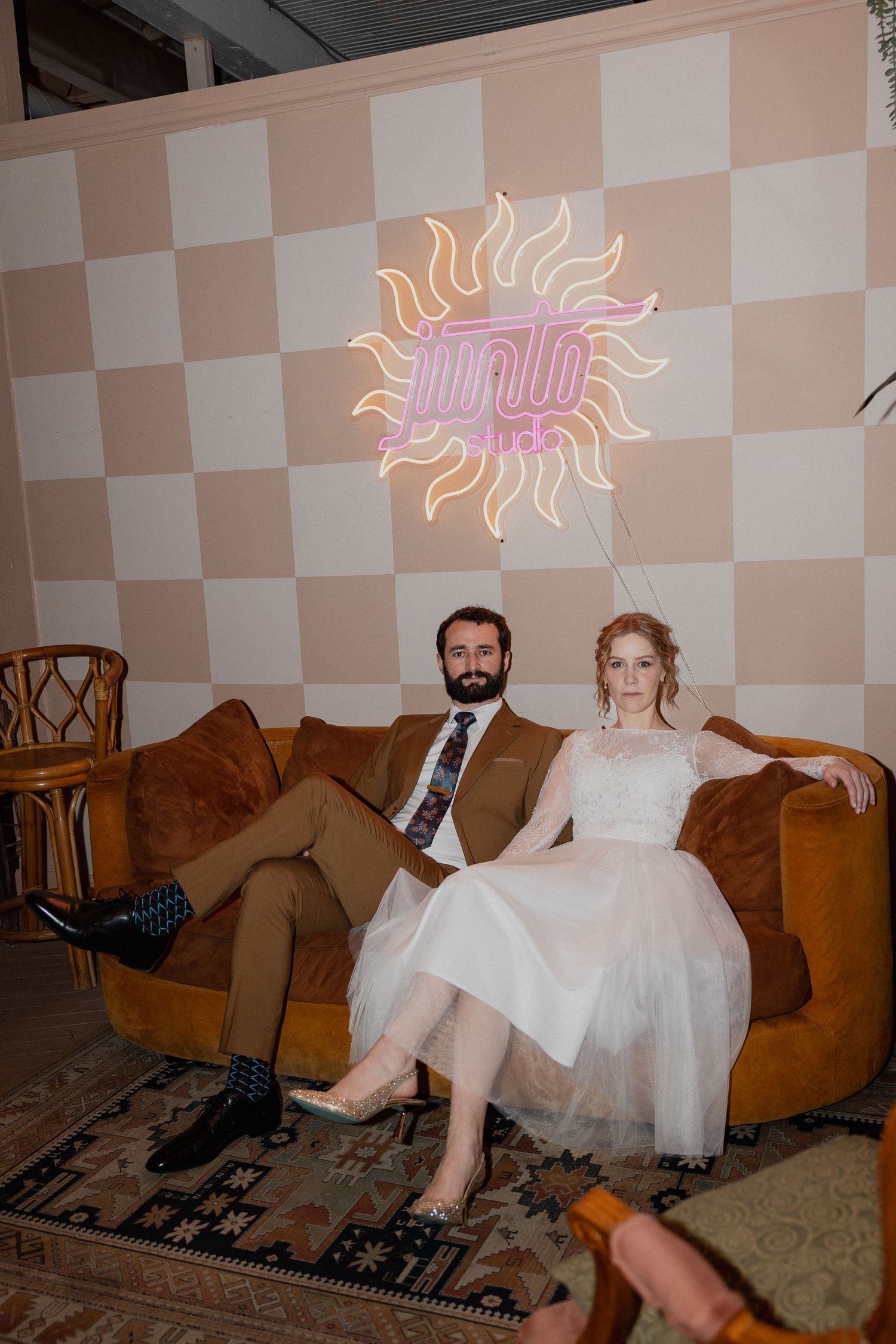 Wedding couple sits on brown velvet couch beneath pink neon sun sign on checkered wall.