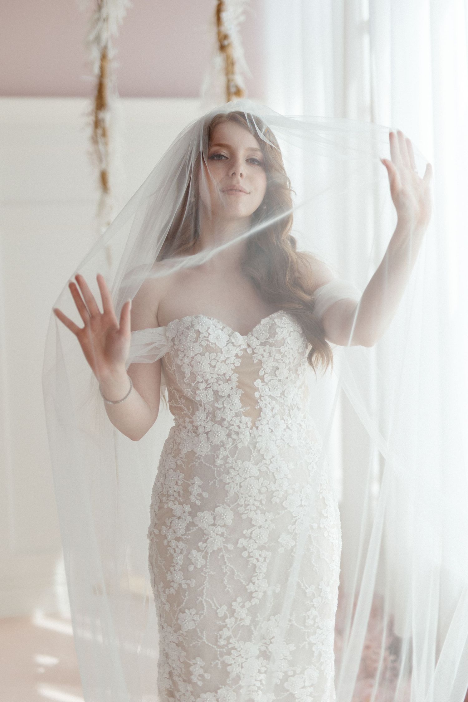 A bride in a detailed lace wedding dress poses behind a sheer veil in soft window light.