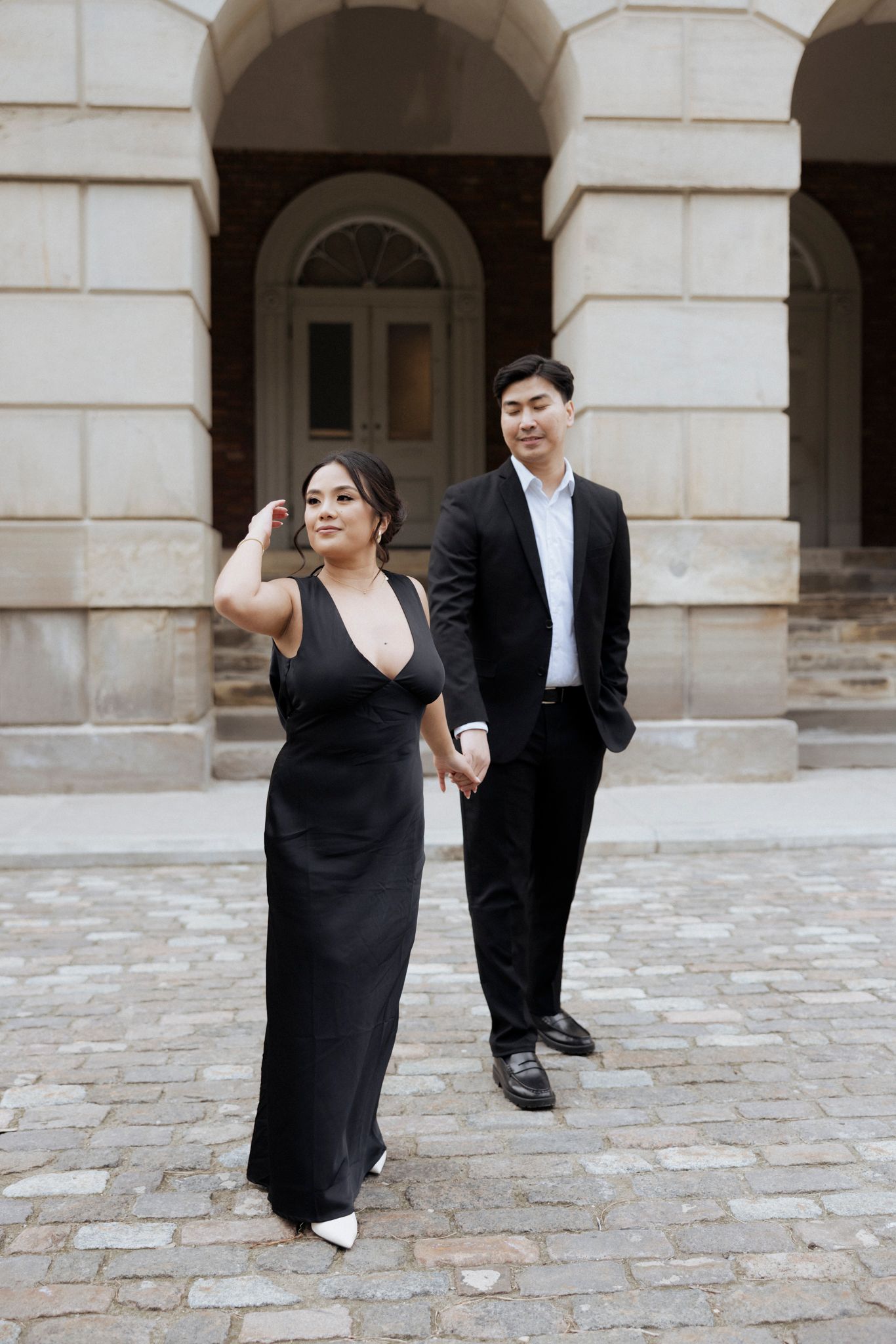 A stylish couple poses in formal wear against classic architecture with stone columns and arches.