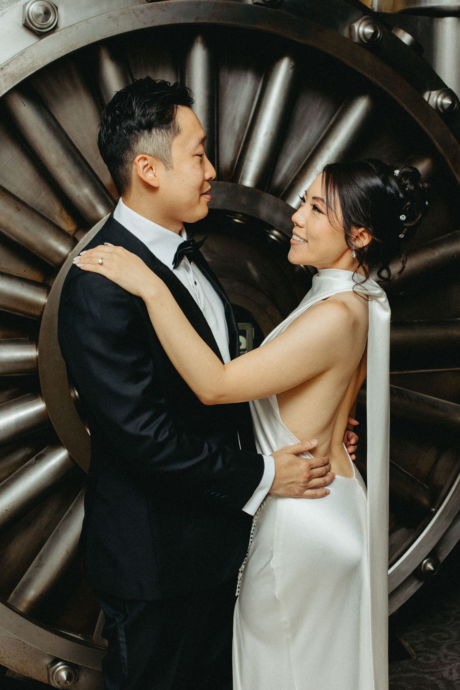 A couple in wedding attire embrace in front of a large jet engine turbine for a dramatic wedding photo.
