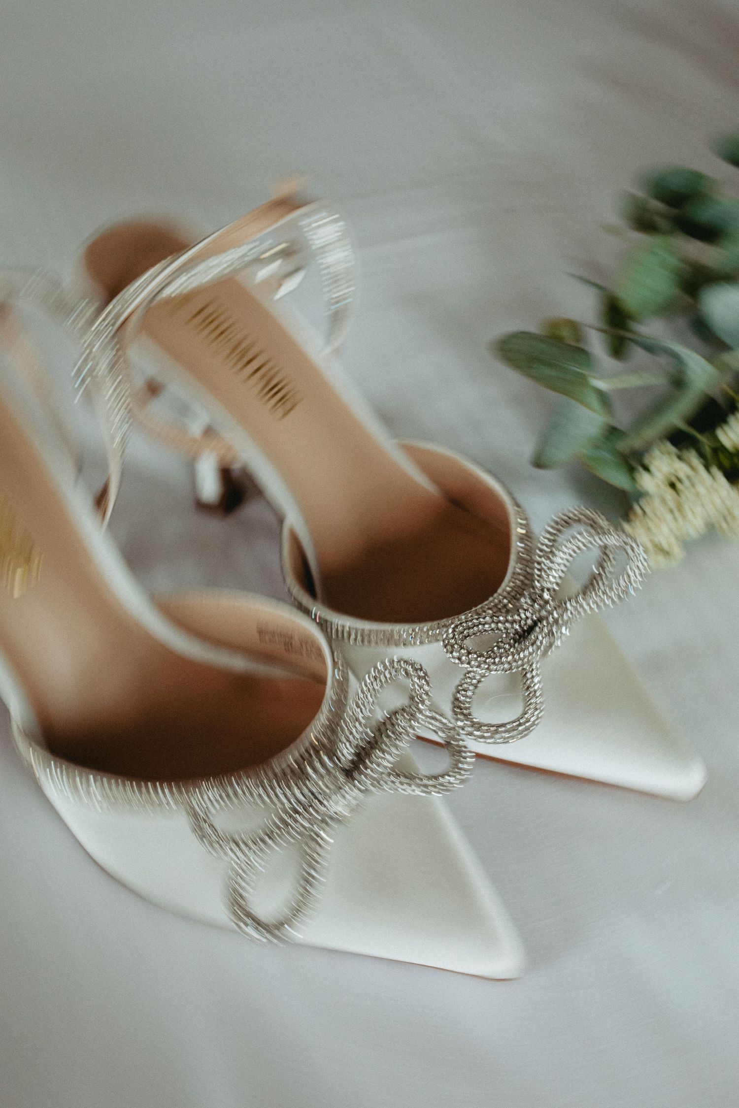 Elegant white bridal shoes with crystal embellishments and stiletto heels displayed beside green foliage.
