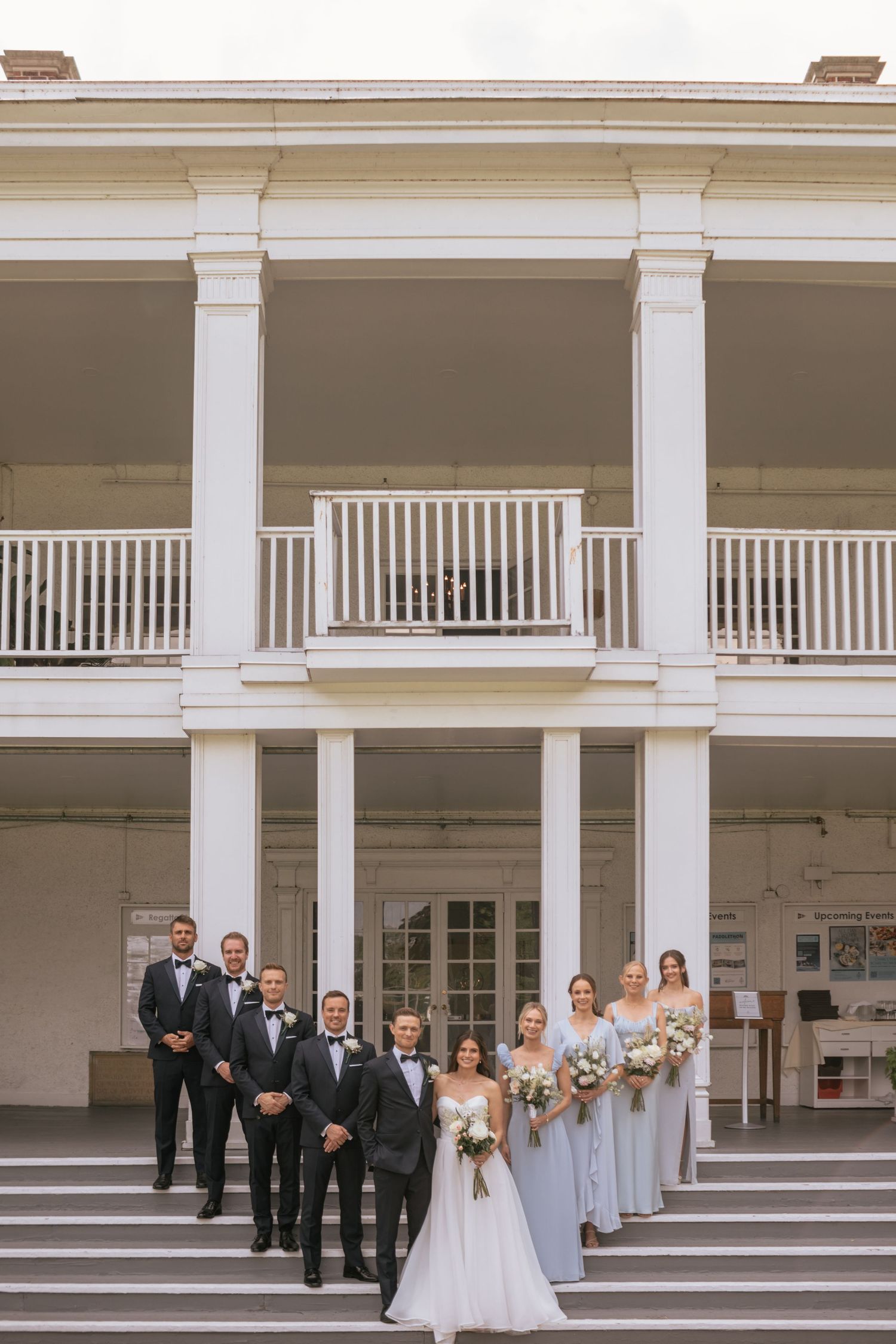 Wedding party stands on grand white columned porch stairs of historic southern style mansion.