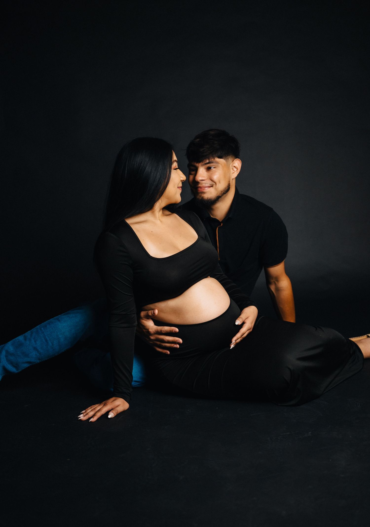 Intimate maternity photo of couple in black clothing against dark background.