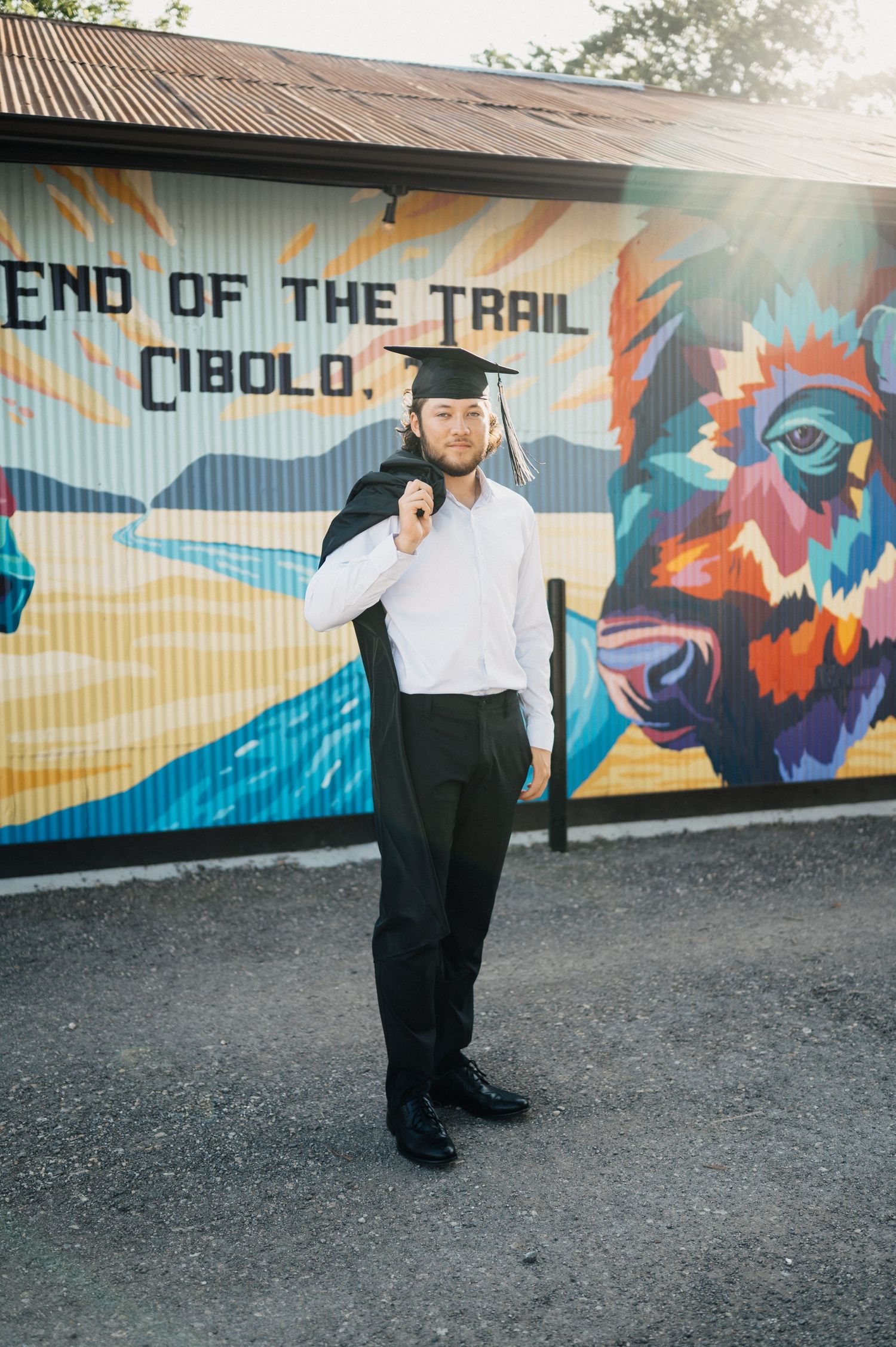 Graduate stands confidently in front of End of the Trail Cibolo mural wearing cap and gown.