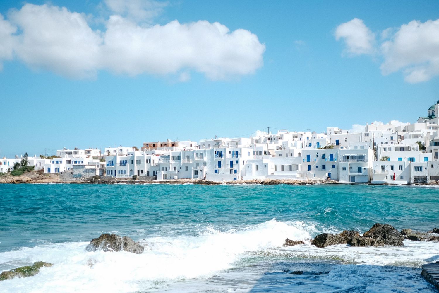 White coastal town buildings overlooking bright blue Mediterranean waters with waves.