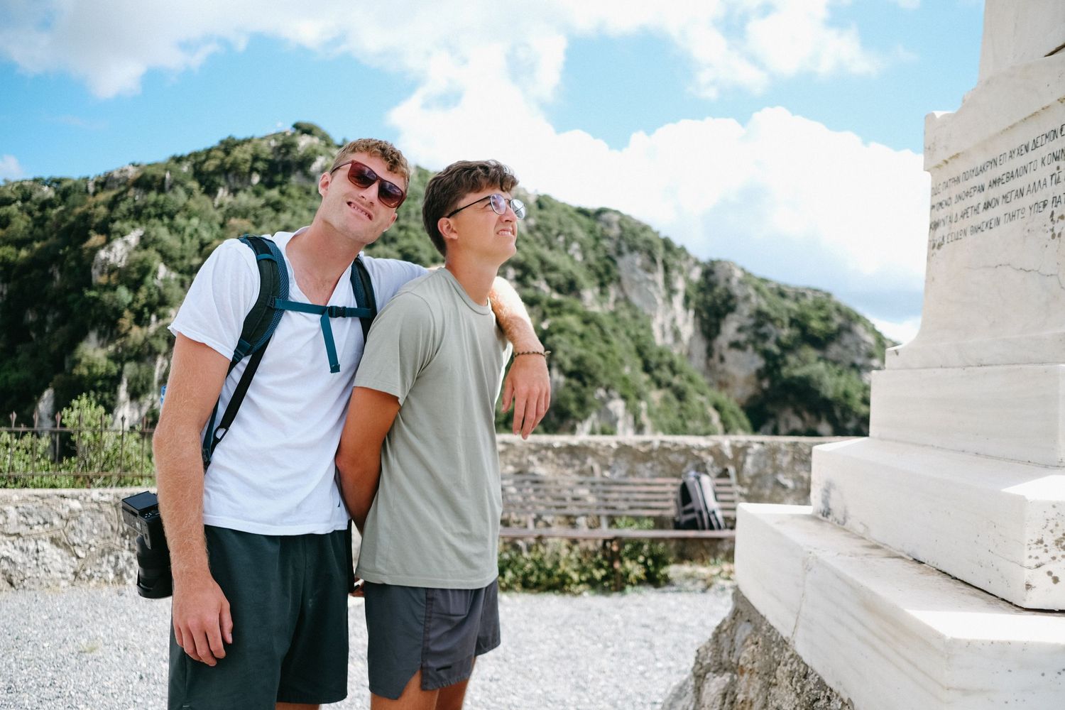 Two travelers posing by a stone monument against a mountainous Mediterranean landscape.