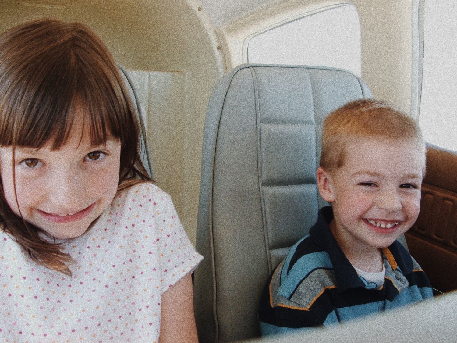 Two children smile while sitting in airplane seats during flight.