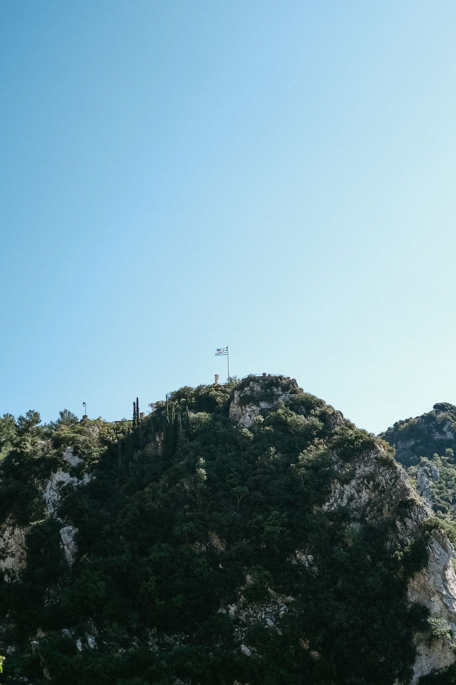 A steep rocky mountain peak with a flag at the summit against a bright blue sky.