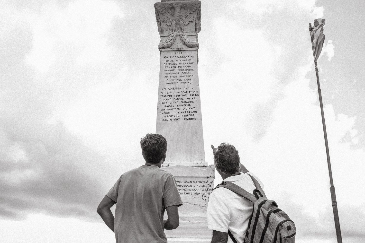 Two people viewing a stone monument with Greek inscription and flag.