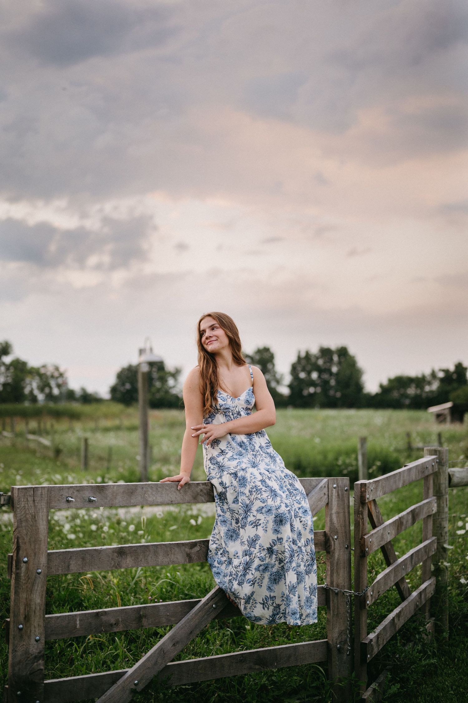 Woman in blue floral dress sits peacefully on rustic wooden fence in countryside meadow at sunset.