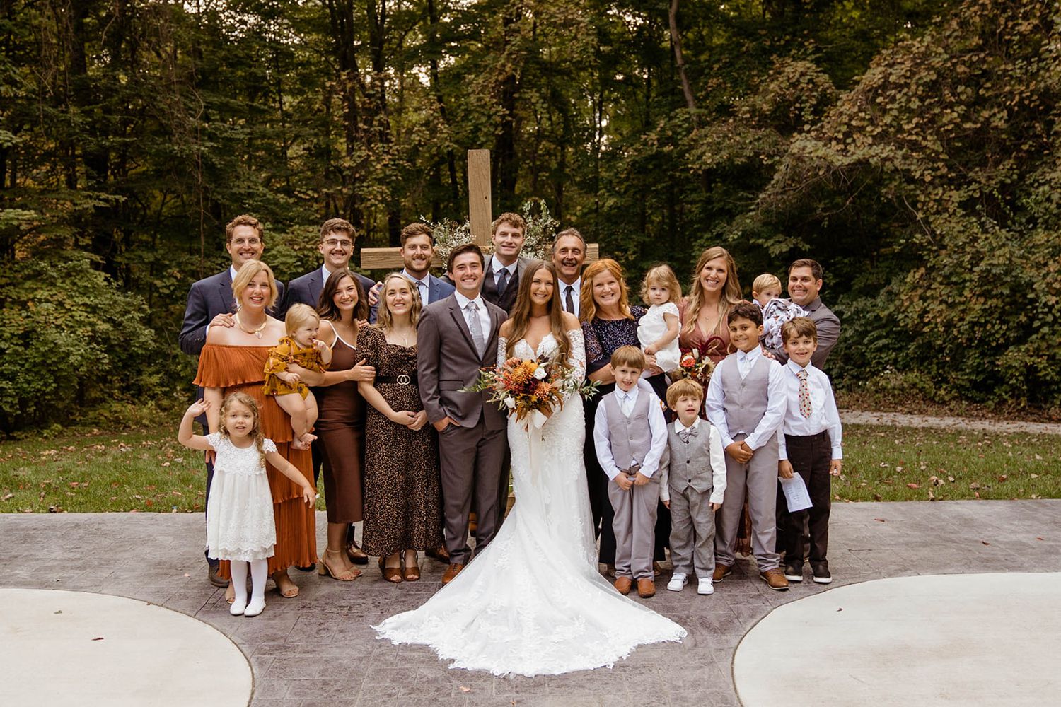 A large wedding party poses outdoors in coordinated brown and gray attire with autumn foliage behind them.