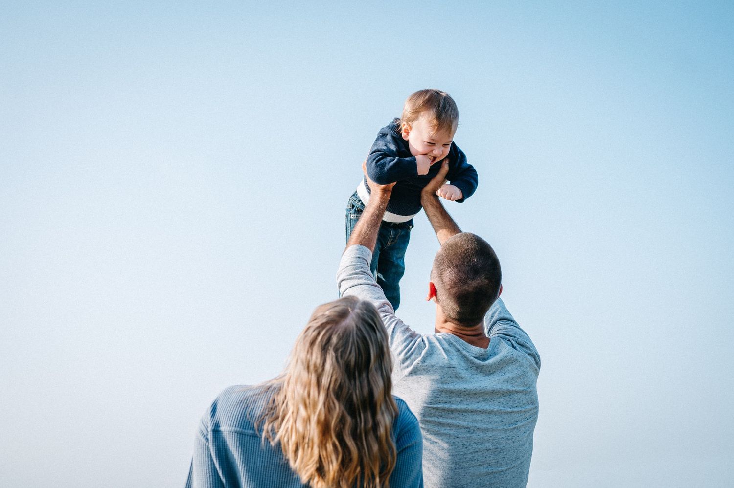 Parents playfully lifting baby against blue sky background during outdoor family moment.