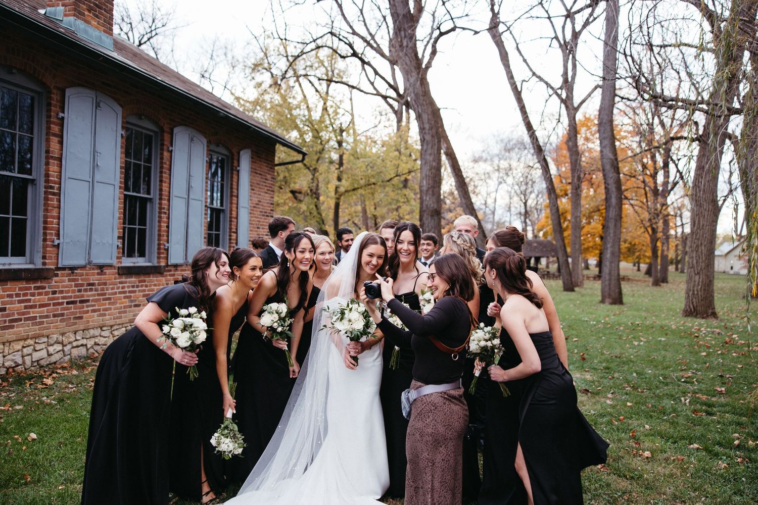 Wedding party in black dresses poses with bride outside a rustic brick building in autumn.