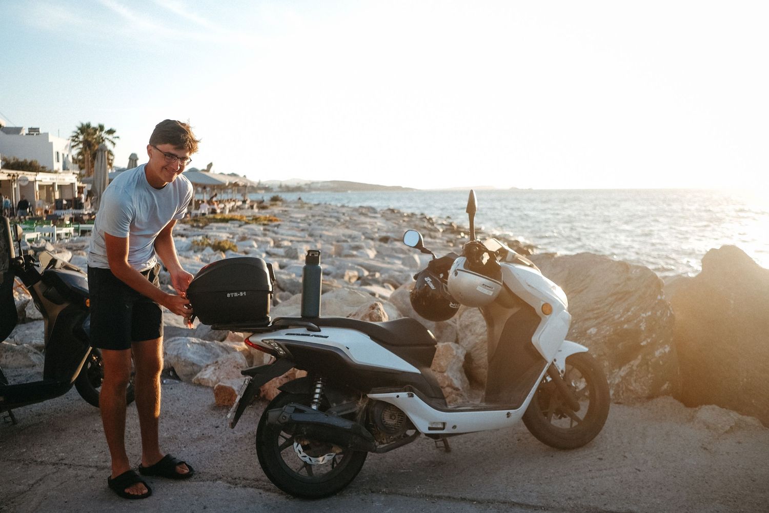 Person examining a parked scooter near coastal rocks at sunset.