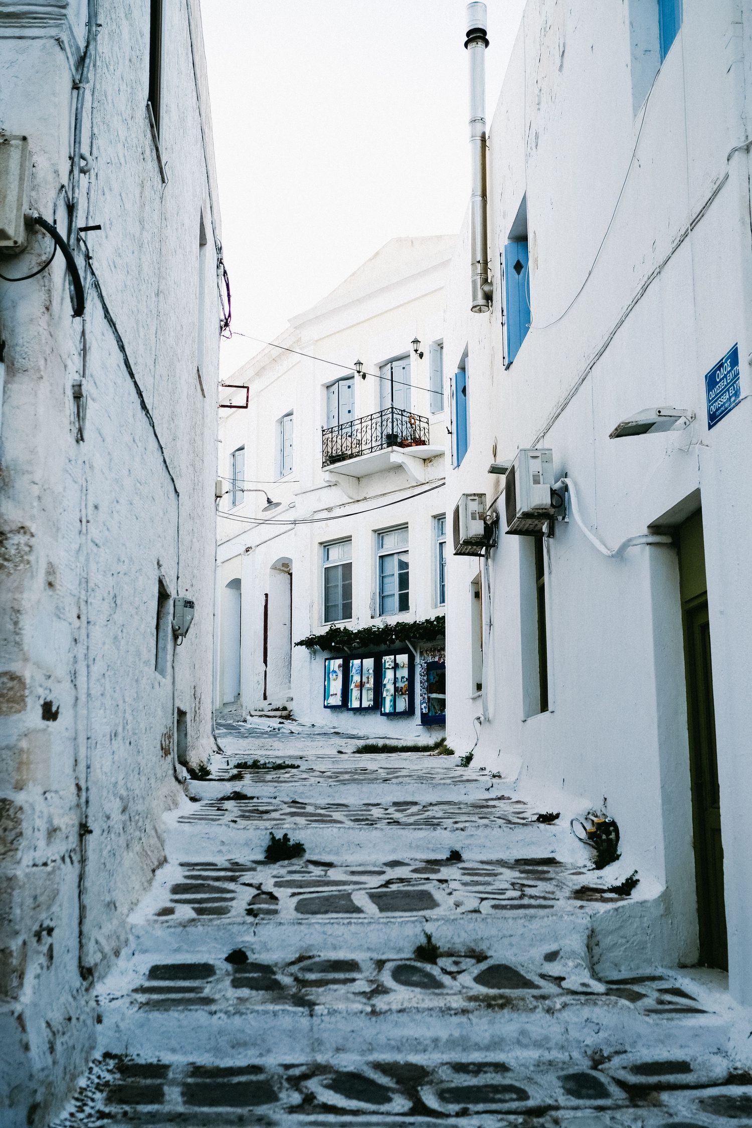 Empty cobblestone alley between white buildings with blue accents and balconies.