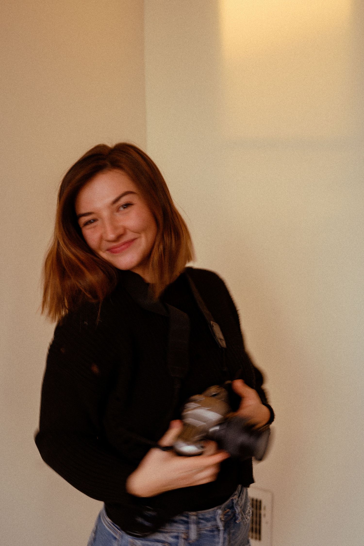Person in black sweater holding something while smiling in warm indoor lighting.