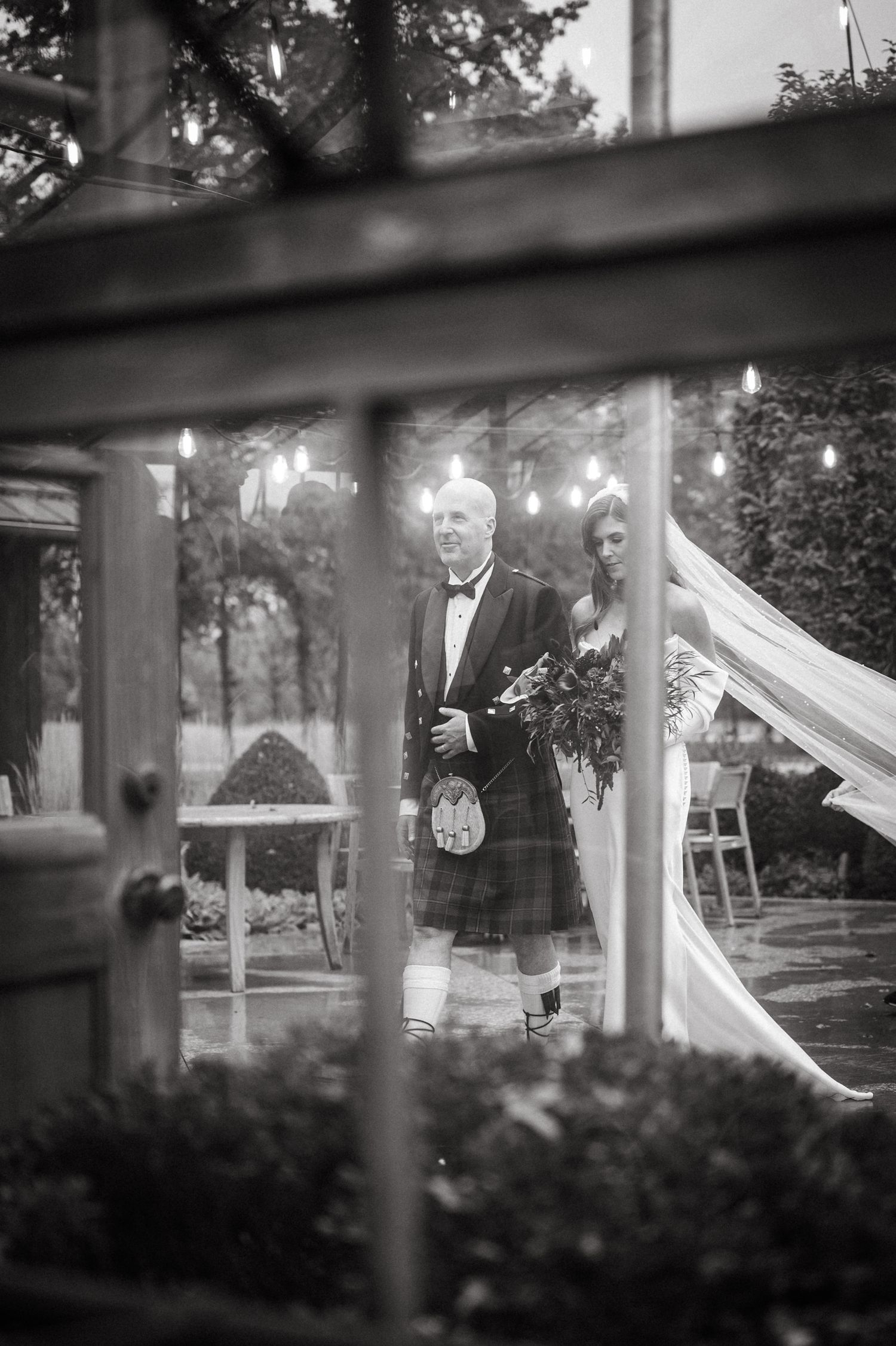 A traditional Scottish groom in a kilt and tuxedo jacket stands under an outdoor wedding tent.