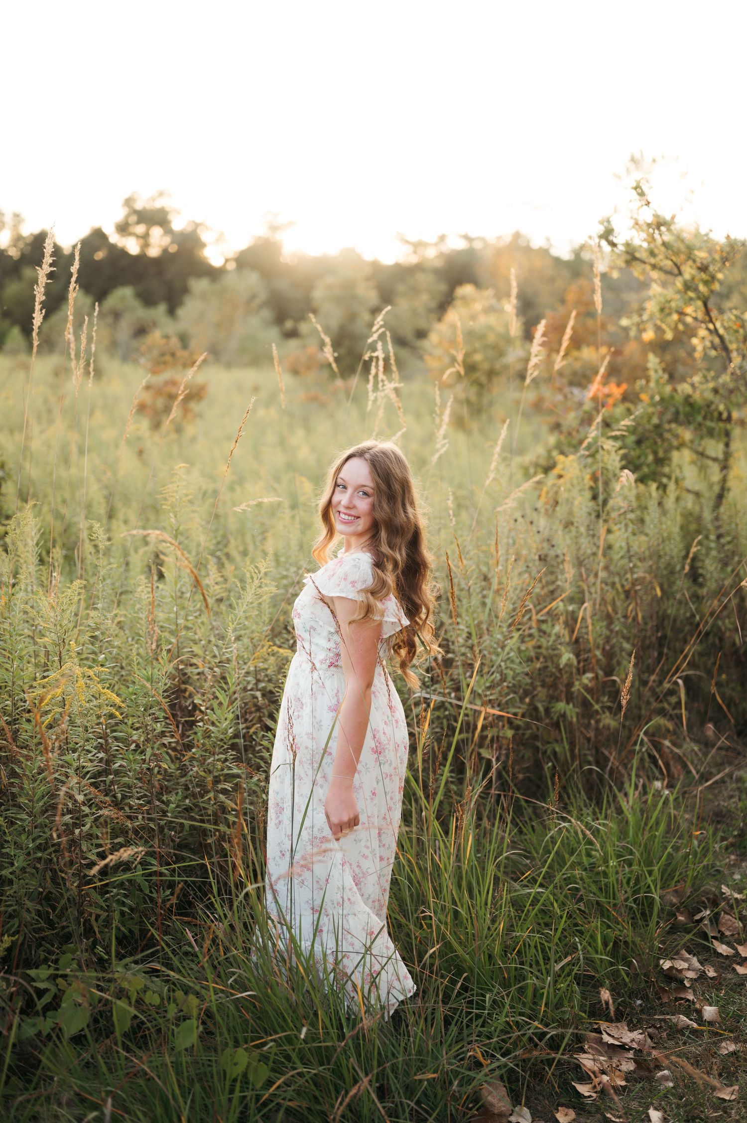 A person in a white lace dress stands in a sunlit meadow surrounded by tall grass and wildflowers at golden hour.