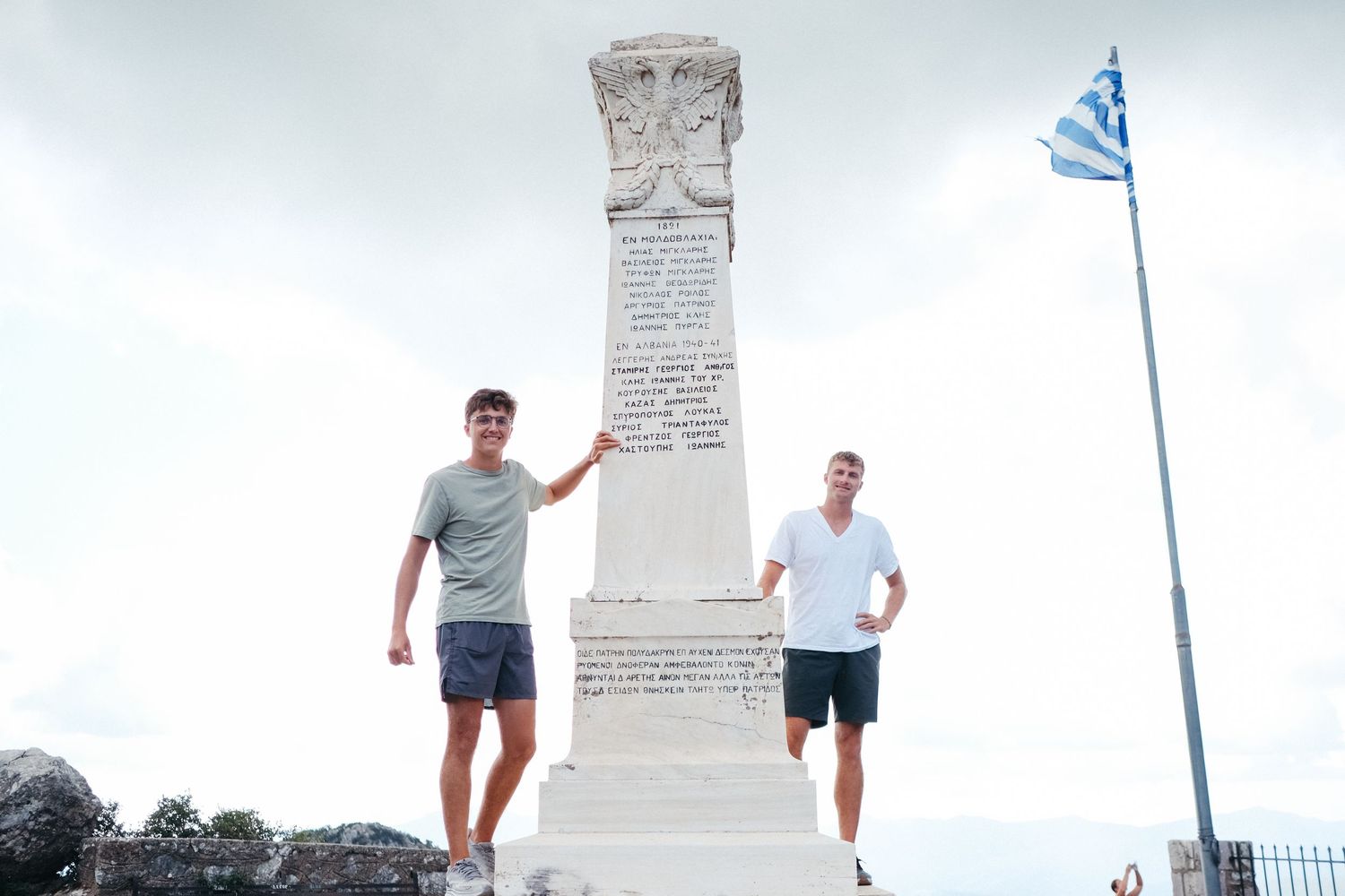 Two visitors standing beside a memorial monument with a Greek flag waving in the background.