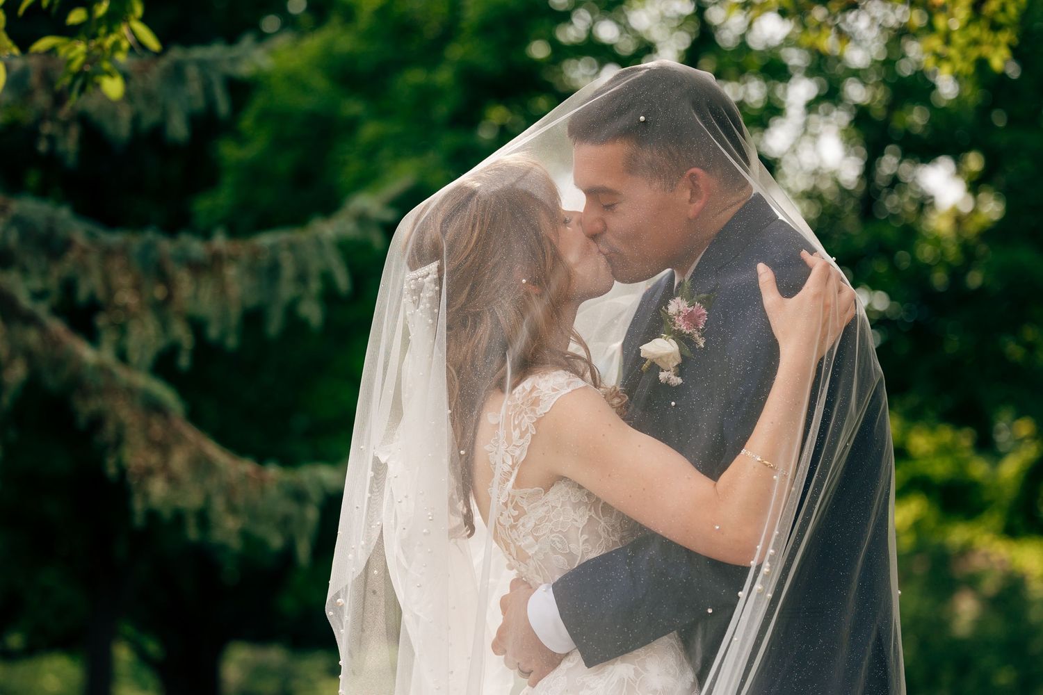 Bride and groom share a romantic kiss under a sheer wedding veil, surrounded by lush green trees at an outdoor wedding.
